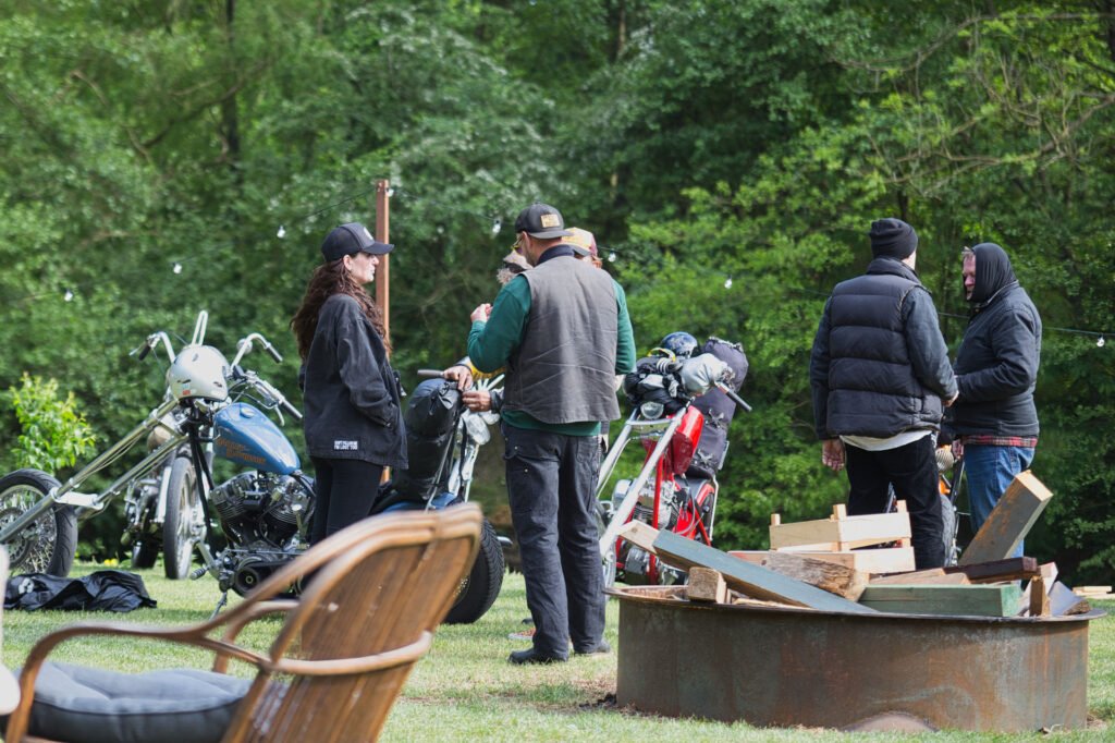 Eine Gruppe von Menschen in Freizeitkleidung steht und unterhält sich in der Nähe von Motorrädern und einer Feuerstelle mit Holz, im Freien auf einer Wiese mit Bäumen im Hintergrund. Im Vordergrund ist ein leerer Stuhl zu sehen.