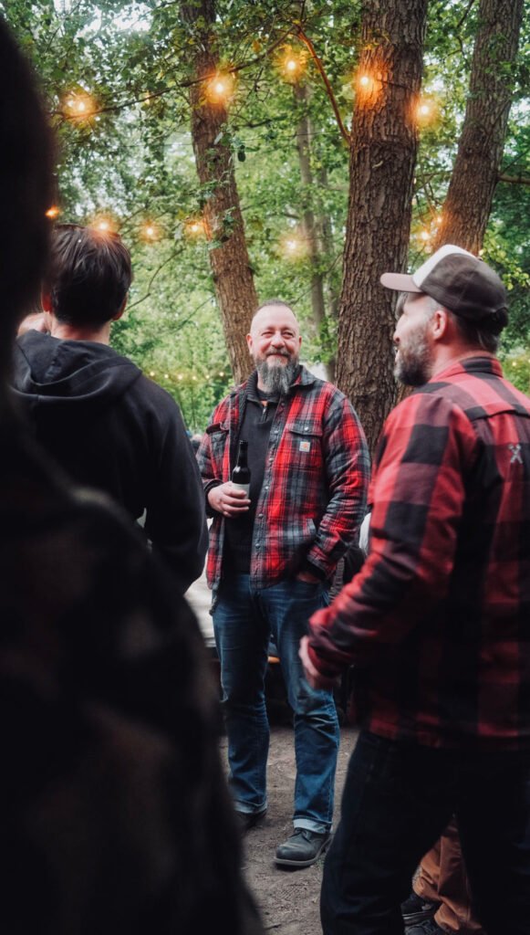 Eine Gruppe von Menschen in karierten Hemden steht im Freien unter Lichterketten in einem Waldstück. Ein Mann mit Bart und einem Getränk lächelt den anderen in der Gruppe freundlich zu.