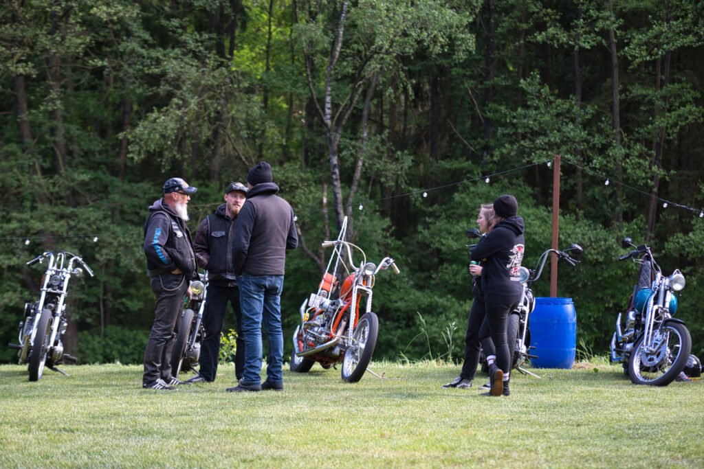 Vier Personen unterhalten sich in der Nähe von Custom-Motorrädern auf einer von Bäumen umgebenen Wiese. Zwei Männer tragen schwarze Jacken und Mützen, während zwei Frauen, ebenfalls in dunkler Kleidung, in der Nähe stehen. Lichterketten und ein blaues Fass sind zu sehen.