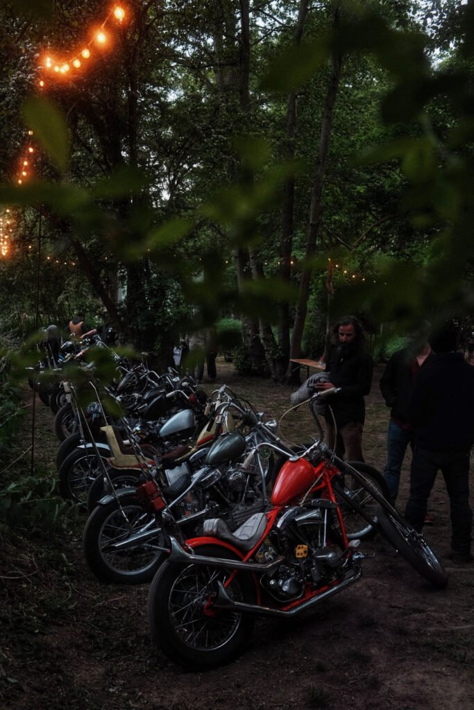 Eine Reihe geparkter Motorräder, darunter ein auffälliges rotes Motorrad, säumen einen Feldweg in einem Waldgebiet. In der Nähe stehen Menschen zwischen Bäumen, über denen Lichterketten hängen, die eine gemütliche Atmosphäre im Freien schaffen.