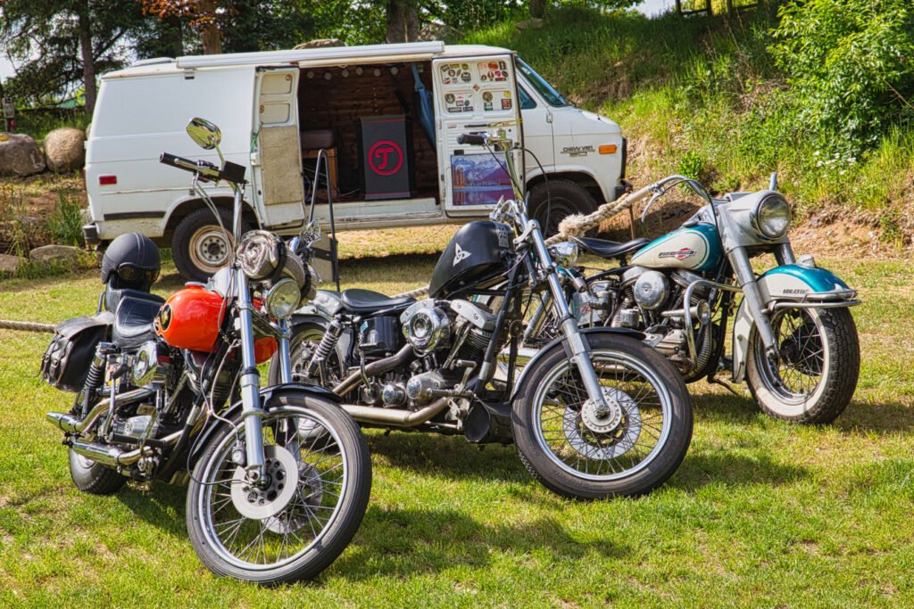 Drei Oldtimer-Motorräder sind auf dem Rasen vor einem klassischen weißen Lieferwagen geparkt, dessen Seitentür geöffnet ist und einen gemütlichen, mit Aufklebern und Postern dekorierten Innenraum offenbart. Im Hintergrund sind Bäume und Grünflächen zu sehen.