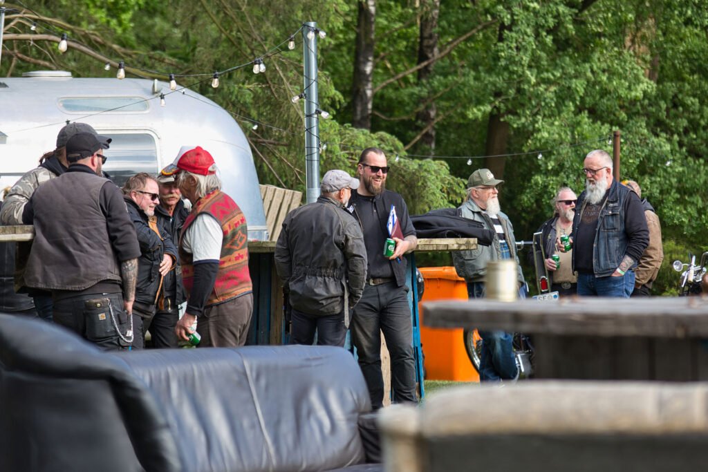 Eine Gruppe von Menschen in legerer Kleidung und im Bikerstil versammelt sich im Freien in der Nähe eines glänzenden silbernen Wohnwagens, plaudert und hält Getränke in der Hand, während im Hintergrund Bäume und Lichterketten zu sehen sind.