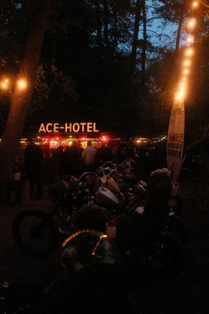 Eine Gruppe von Motorrädern, die in der Abenddämmerung vor einer warm beleuchteten Bar mit einem leuchtenden ACE HOTEL"-Schild geparkt sind, umgeben von Bäumen und Menschen, mit Lichterketten über dem Kopf.