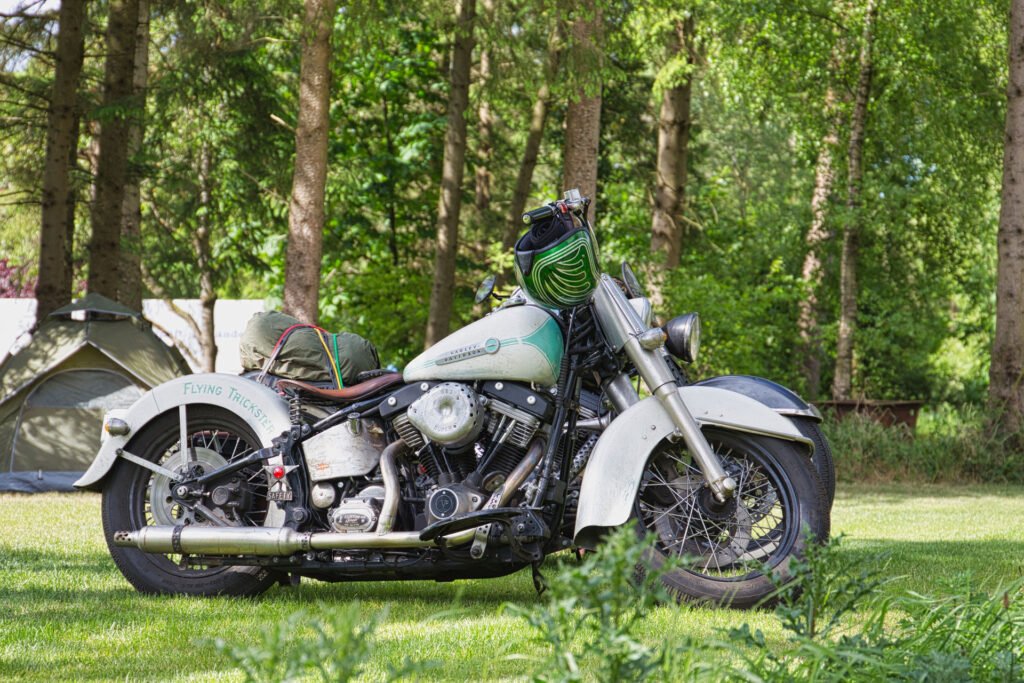 Ein altes weißes Motorrad mit Satteltaschen steht auf einer Wiese in einem Waldgebiet in der Nähe eines grünen Zeltes, auf dessen Rücken eine Campingausrüstung befestigt ist. Bäume und Grünpflanzen füllen den Hintergrund.