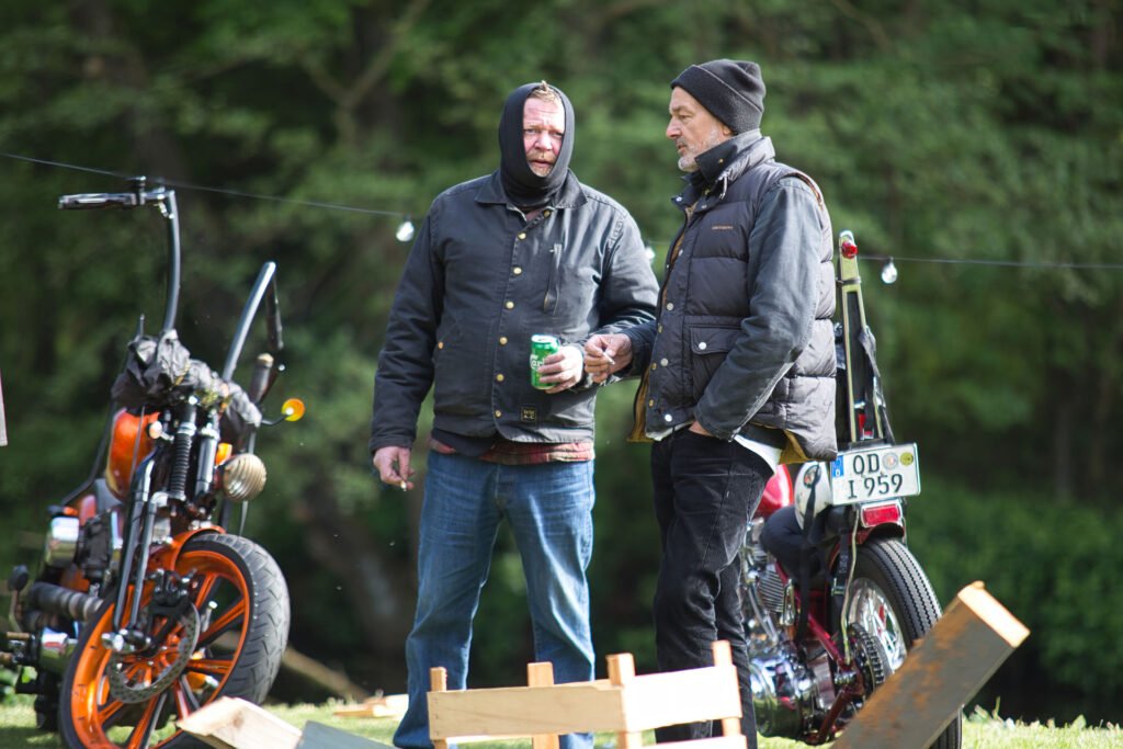 Zwei Männer in warmen Jacken und Mützen stehen draußen neben zwei Motorrädern und unterhalten sich. Einer hält eine Getränkedose in der Hand. Holzbretter und Lichterketten sind zu sehen, im Hintergrund grüne Bäume.