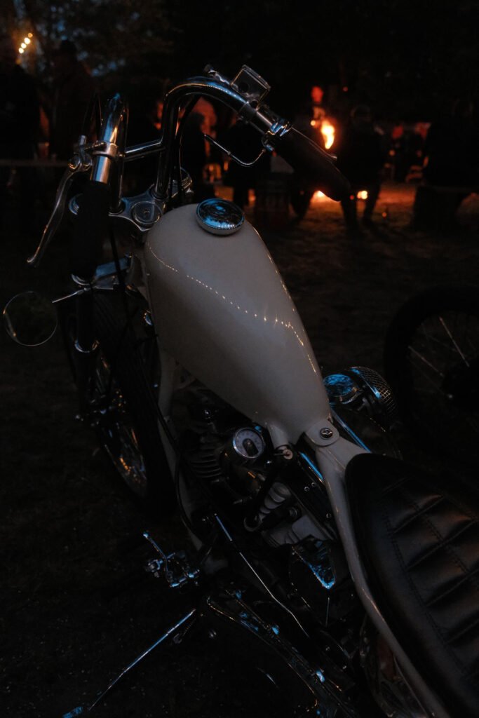Nahaufnahme eines klassischen Motorrads mit weißem Tank bei Nacht, mit einer Gruppe von Menschen und einem glühenden Lagerfeuer im Hintergrund.