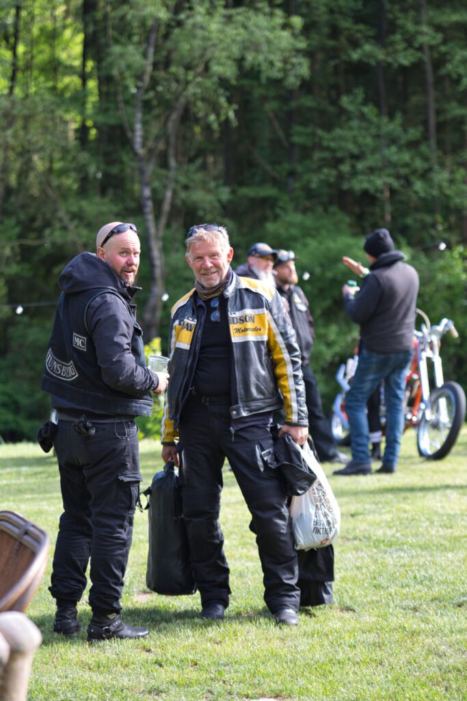 Zwei Männer in schwarzer Motorradkleidung stehen lächelnd im Gras und halten Taschen in der Hand. Weitere Personen und ein Motorrad befinden sich im Hintergrund in der Nähe von Bäumen. Die Szene wirkt ungezwungen und im Freien.