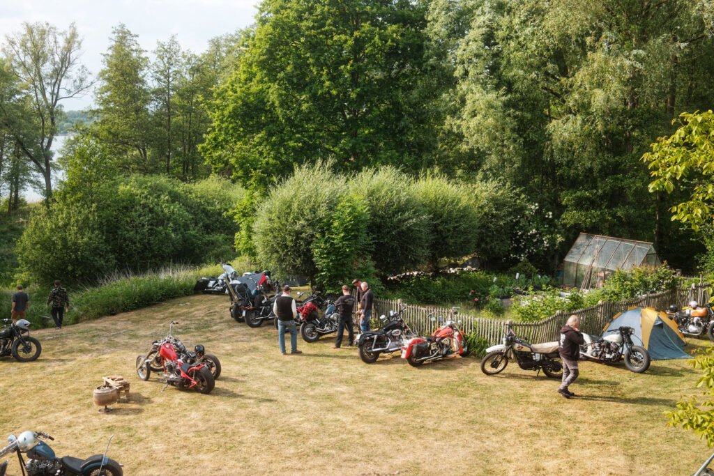 Eine Gruppe von Personen mit mehreren Motorrädern versammelt sich auf einer von Bäumen umgebenen Wiese in der Nähe eines Zeltes und eines Gewächshauses, wobei einige Personen stehen und andere auf Fahrrädern sitzen.