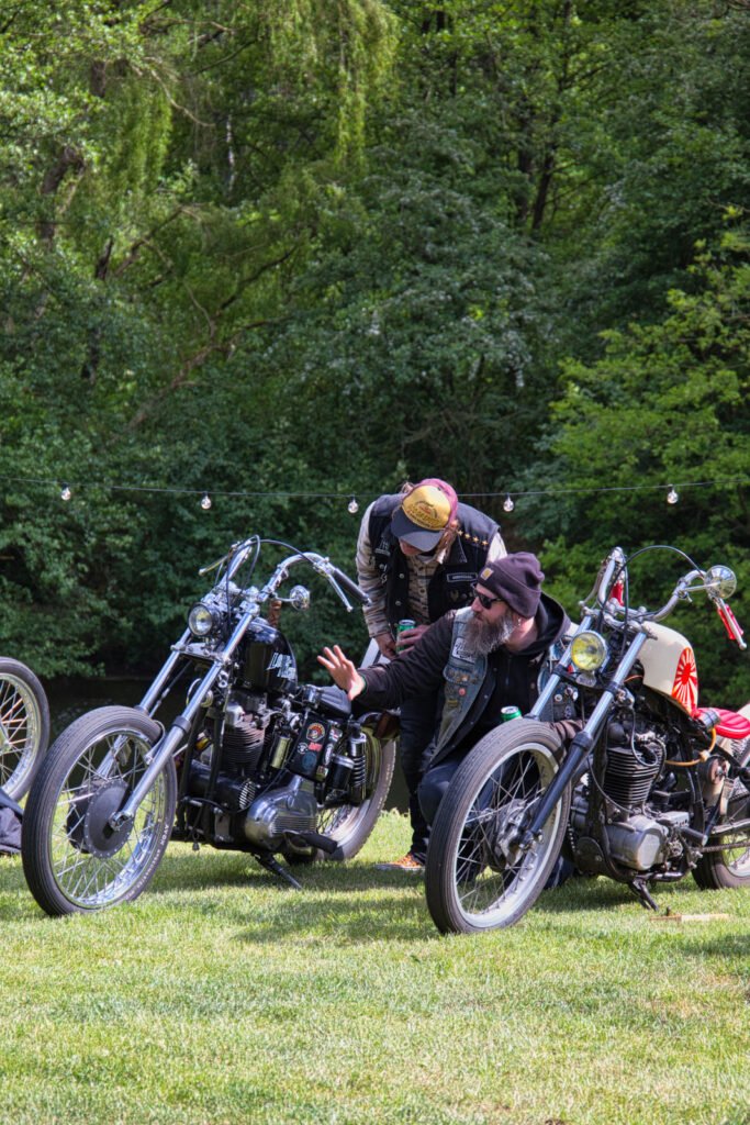 Zwei Männer in legerer Outdoor-Kleidung unterhalten sich neben ihren Oldtimer-Motorrädern auf einer Wiese, umgeben von Bäumen und hängenden Lichterketten im Hintergrund.