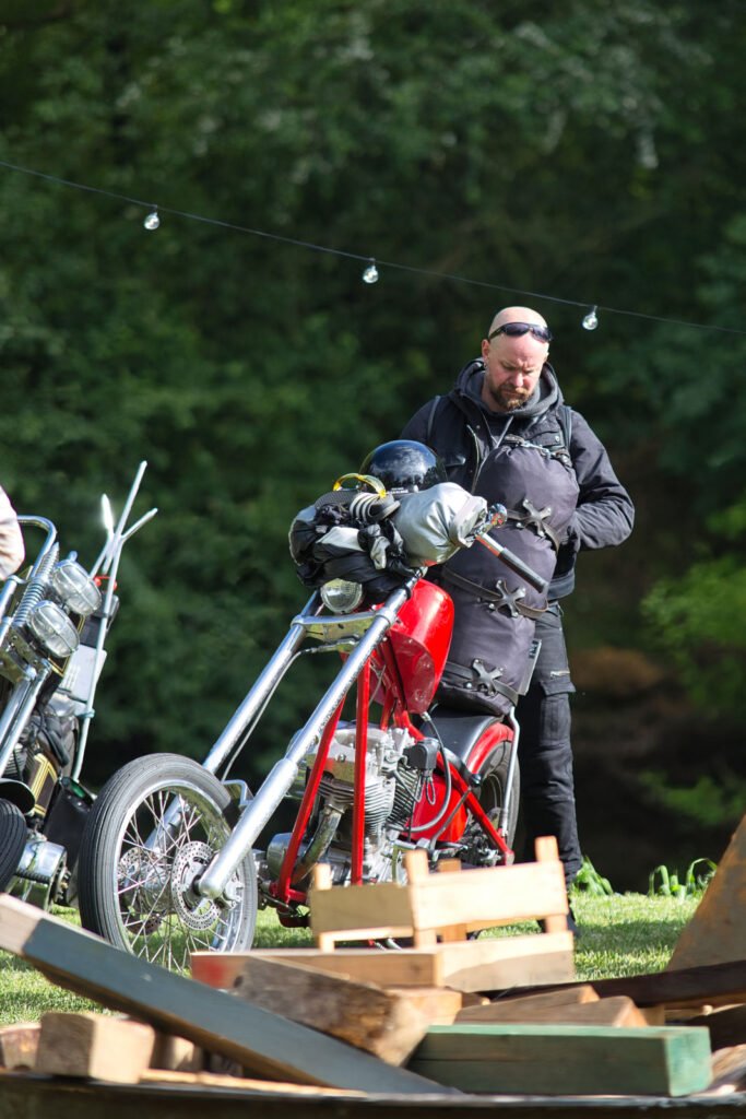 Ein Mann in schwarzer Kleidung und mit Sonnenbrille steht im Freien neben einem roten Motorrad im Chopper-Stil. Im Hintergrund sind Grünflächen zu sehen, im Vordergrund Holzplanken.