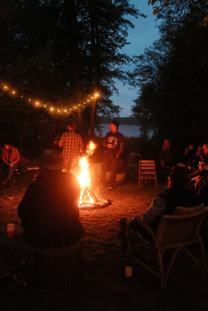 Eine Gruppe von Menschen sitzt und steht um ein helles Lagerfeuer in der Abenddämmerung. Lichterketten hängen über dem Feuer, Bäume umgeben den Platz, und im Hintergrund ist der See zu sehen. Einige Leute unterhalten sich, während andere das Feuer beobachten.