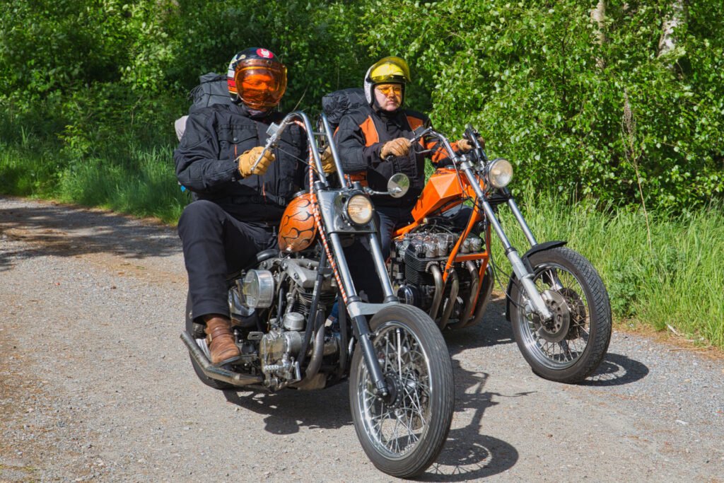 Zwei Personen in schwarzer Kleidung und mit Helmen fahren mit großen Motorrädern auf einer sonnigen, von Bäumen gesäumten Schotterstraße. An den Motorrädern sind Taschen befestigt, und beide Fahrer tragen Sonnenbrillen. Der Hintergrund ist üppig und grün mit Laub.