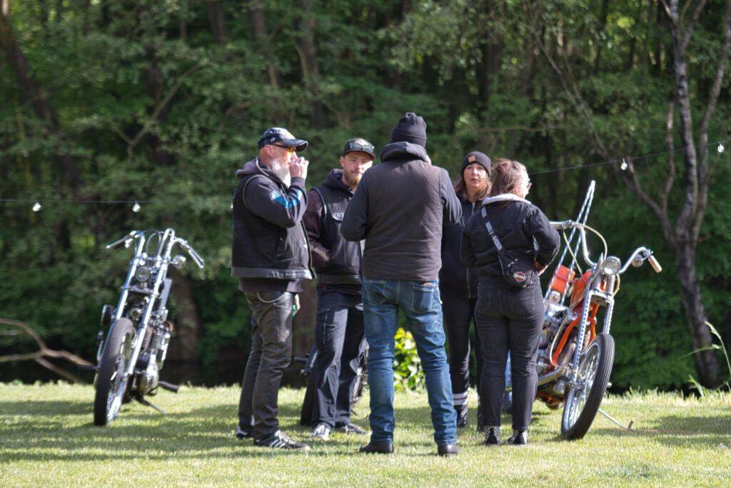 Fünf Personen mit Jacken und Hüten stehen auf einer Wiese in der Nähe von zwei Custom-Motorrädern und unterhalten sich miteinander. Im Hintergrund stehen Bäume, und über ihnen sind Lichterketten zu sehen.