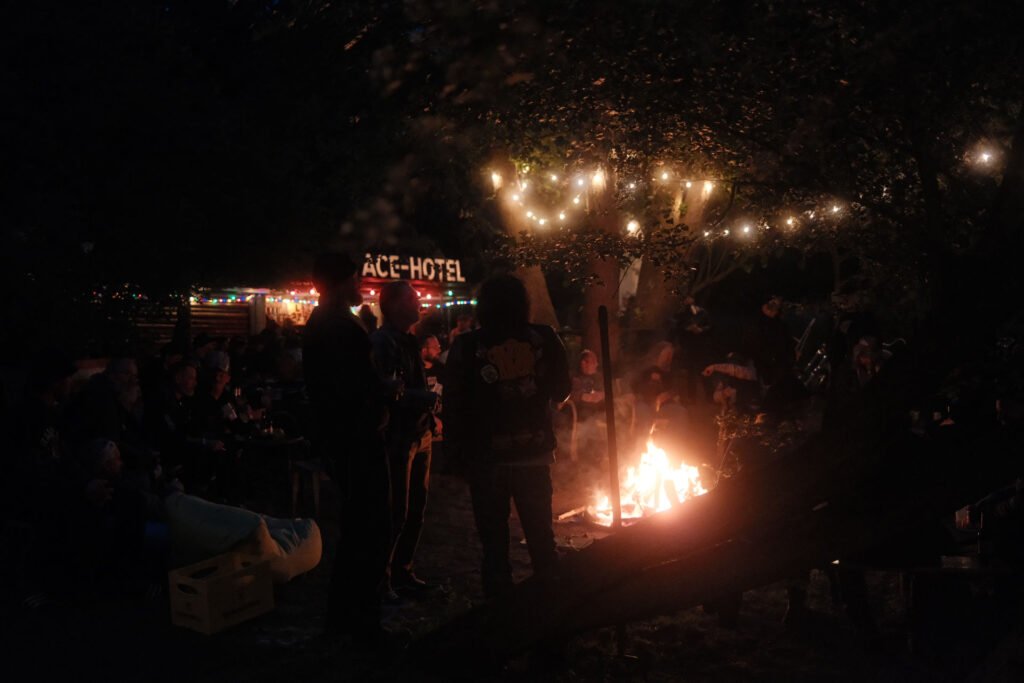 Eine Gruppe von Menschen versammelt sich nachts um ein Lagerfeuer, das von Lichterketten beleuchtet wird, die von Bäumen hängen. Im Hintergrund sind bunte Lichter und ein Schild mit der Aufschrift "ACE HOTEL" zu sehen. Die Atmosphäre wirkt lebendig und gesellig.