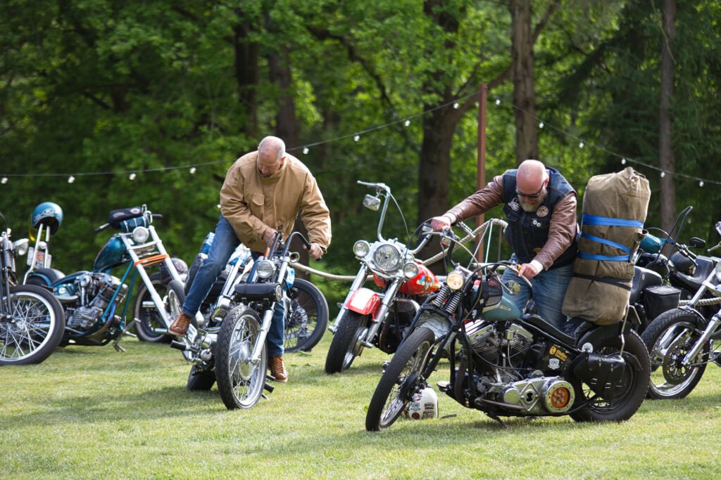 Zwei Personen in Jacken bereiten sich auf eine Fahrt mit einem Oldtimer-Motorrad auf einer Wiese vor, umgeben von anderen geparkten Motorrädern, mit Bäumen und Lichterketten im Hintergrund.