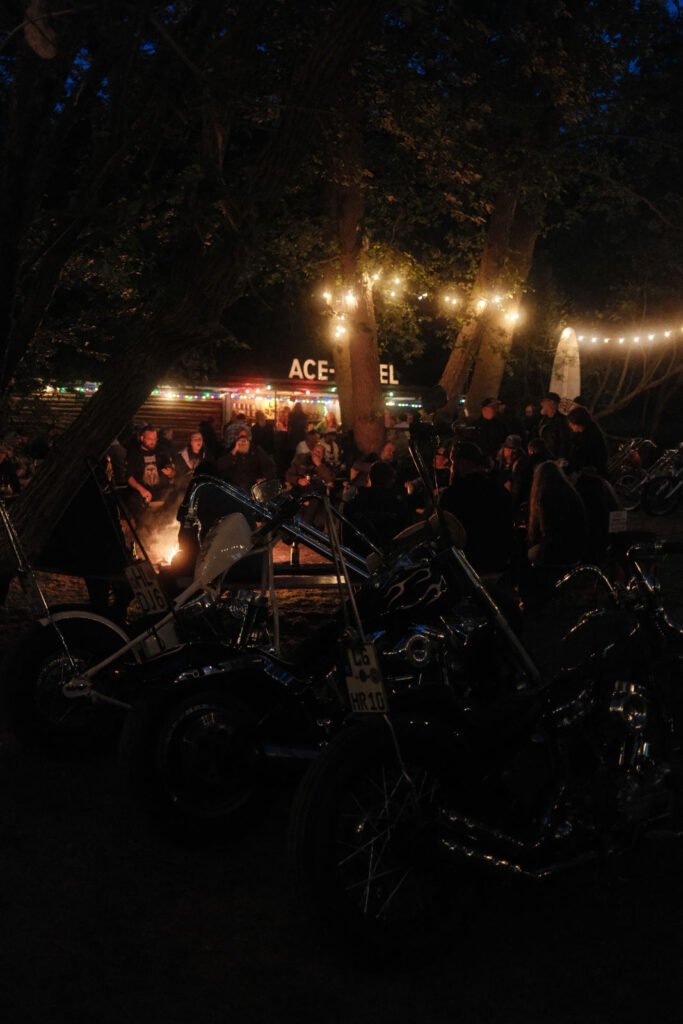 Eine nächtliche Zusammenkunft im Freien mit Menschen, die an einem Lagerfeuer sitzen, im Vordergrund geparkten Motorrädern, an den Bäumen hängenden Lichterketten und einem hell erleuchteten "ACE"-Schild im Hintergrund.