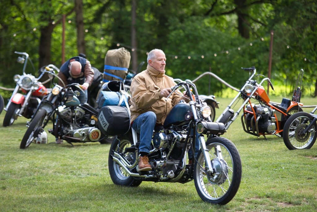 Ein Mann in einer hellbraunen Jacke sitzt auf einem geparkten Motorrad auf einer Wiese mit Bäumen, während ein anderer Mann sein Motorrad in der Nähe einstellt. Im Hintergrund sind mehrere andere Motorräder zu sehen.