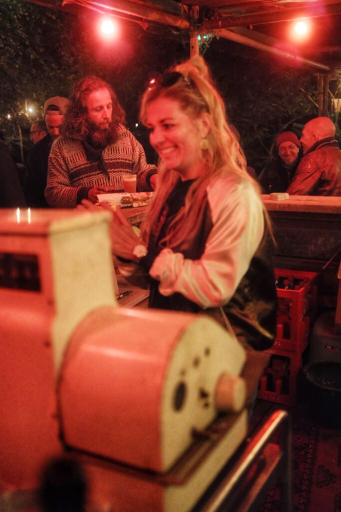 Eine Frau lächelt hinter dem Tresen eines von roten Lichtern beleuchteten Imbissstandes, während ein Mann im Pullover und andere Leute im Hintergrund plaudern. Die Atmosphäre wirkt warm und festlich.