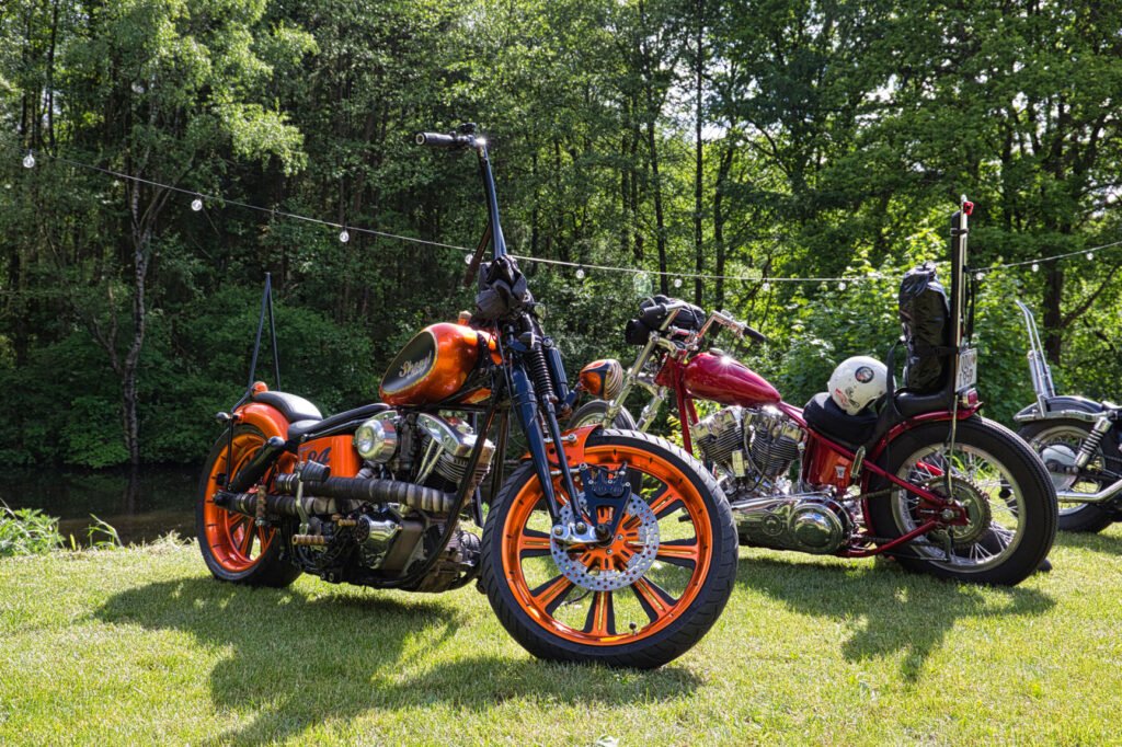 Zwei Custom-Motorräder, ein orange-schwarzes mit orangefarbenen Rädern und ein rotes, verchromtes, sind auf dem Rasen vor Bäumen geparkt. Im Hintergrund hängen Lichterketten. Auf einem der Motorräder befindet sich ein weißer Helm.