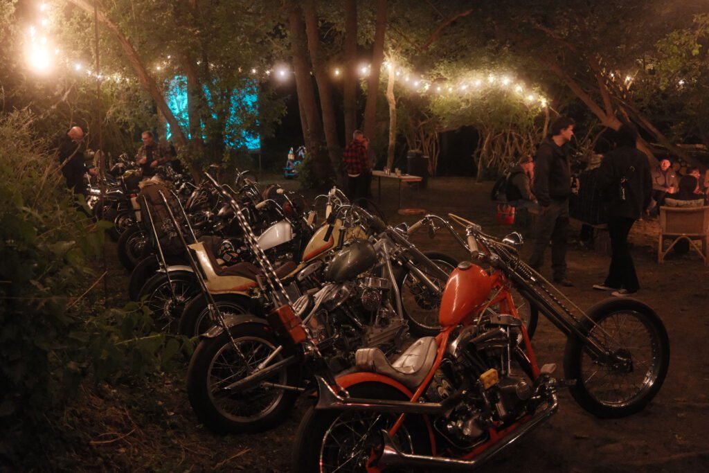 Eine Reihe von Motorrädern ist nachts unter Lichterketten in einem Waldgebiet geparkt, während im Hintergrund Menschen herumstehen und -sitzen und sich unterhalten.