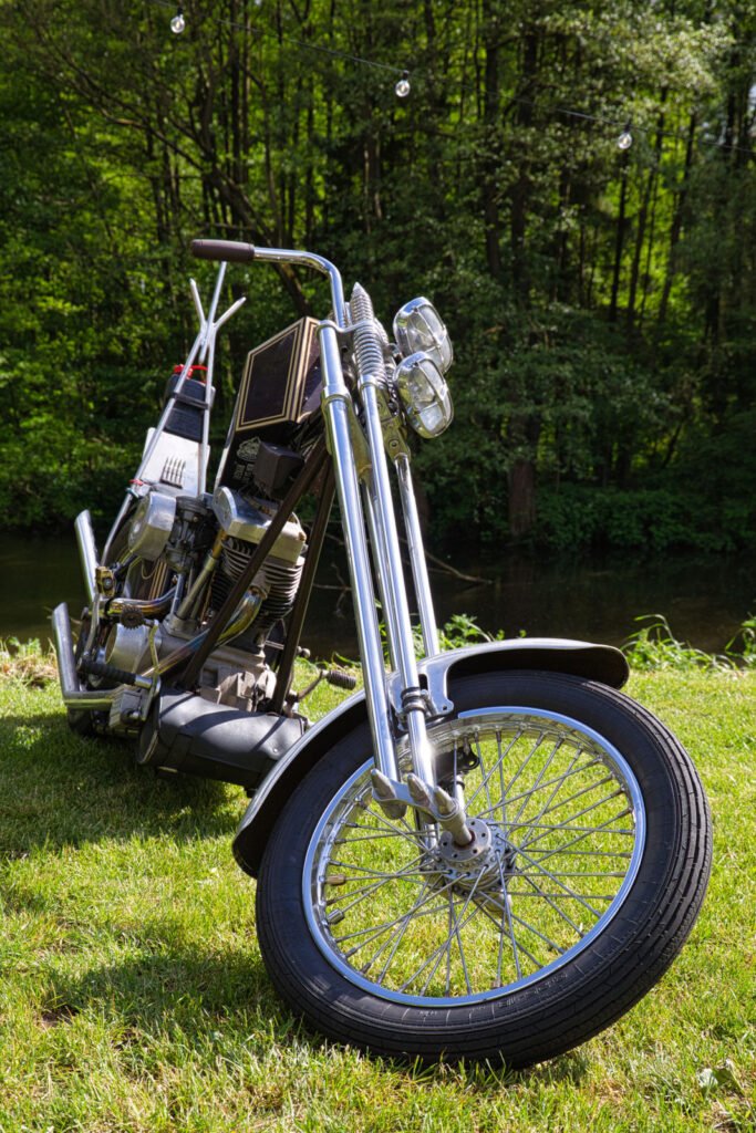 Ein Oldtimer-Motorrad im Chopper-Stil mit verchromten Details ist auf grünem Gras in der Nähe eines Waldgebiets geparkt, mit Bäumen und einem Fluss im Hintergrund.