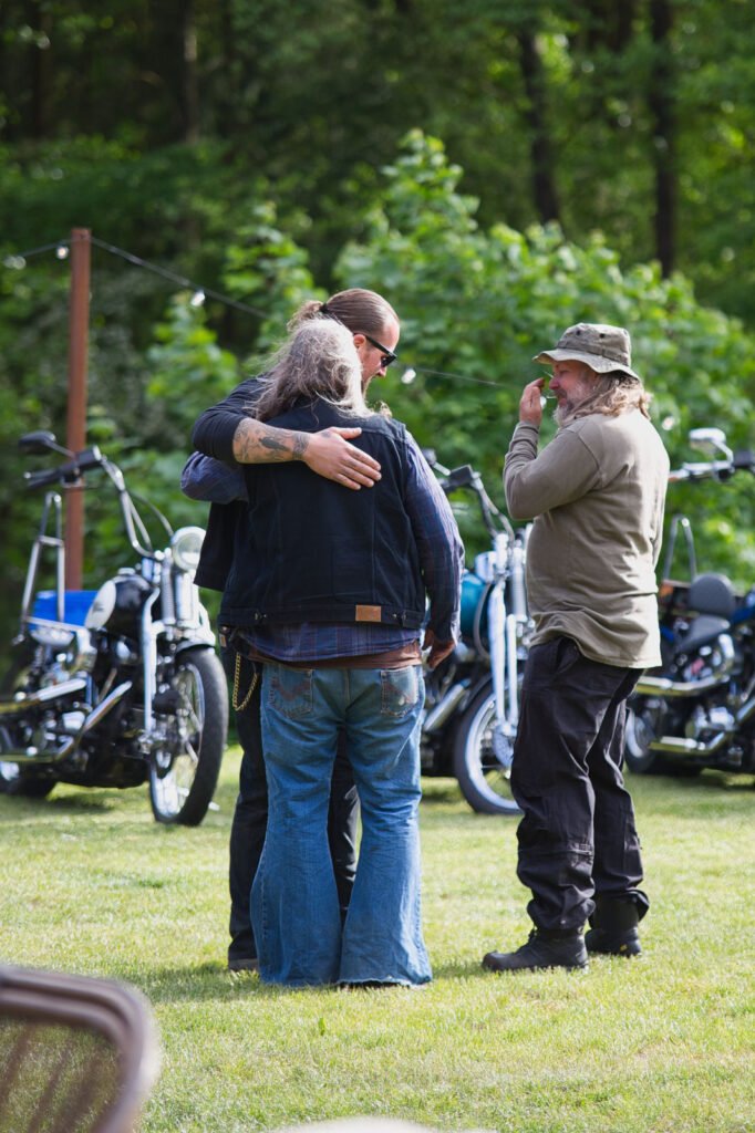 Drei Personen stehen im Freien neben geparkten Motorrädern. Zwei Personen, eine mit langen Haaren, umarmen sich, während eine dritte, bärtige Person mit Hut, lacht und gestikuliert. Grüne Bäume und Gras füllen den Hintergrund, was auf ein zwangloses Beisammensein hindeutet.