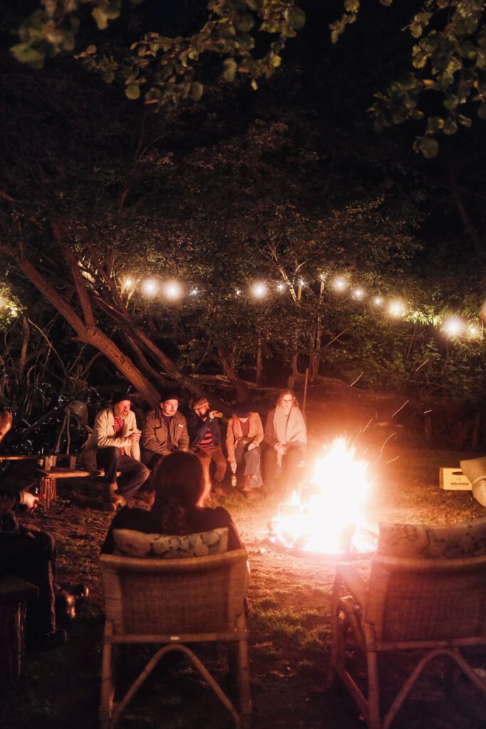 Eine Gruppe von Menschen sitzt nachts um ein glühendes Lagerfeuer, umgeben von Bäumen und Lichterketten. Einige sitzen auf Korbstühlen, andere auf Bänken und genießen ein gemütliches Beisammensein im Freien.