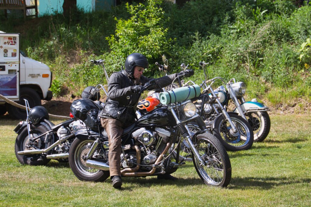Eine Person mit schwarzem Helm und Lederjacke sitzt rittlings auf einem schwarzen Motorrad mit Flammenaufklebern, das zusammen mit zwei anderen Motorrädern auf einer Wiese abgestellt ist. Im Hintergrund sind Bäume und ein weißes Fahrzeug zu sehen.