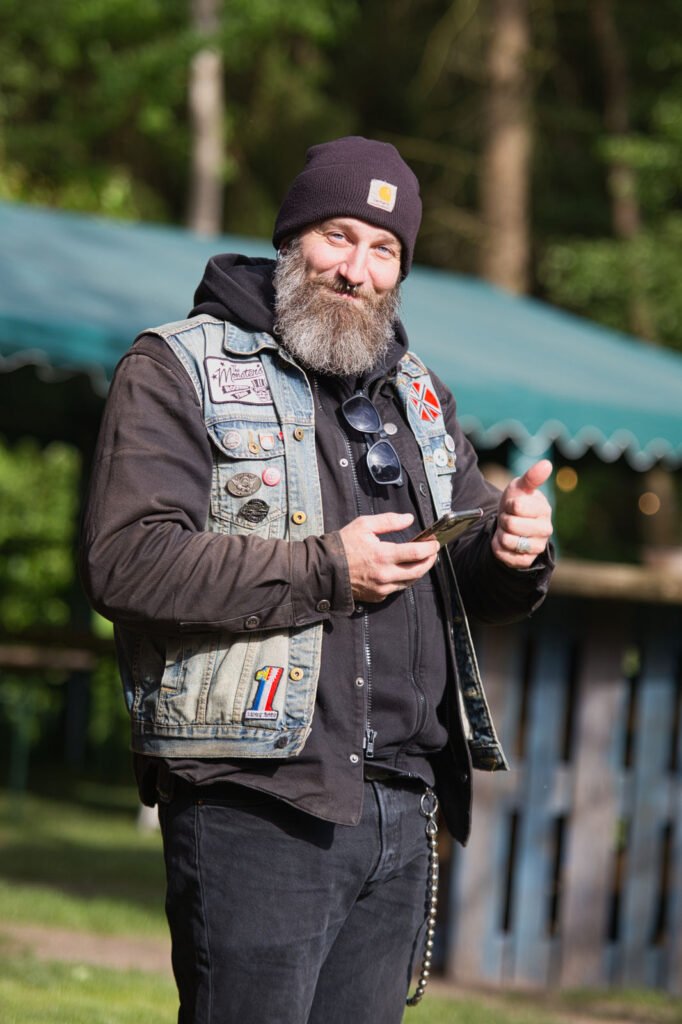 Ein bärtiger Mann mit einer schwarzen Mütze, einem schwarzen Kapuzenpulli und einer Jeansweste mit Aufnähern steht im Freien, lächelt und hält ein Smartphone in der Hand. Er gestikuliert mit seiner linken Hand, im Hintergrund sind grüne Bäume und ein Zelt zu sehen.