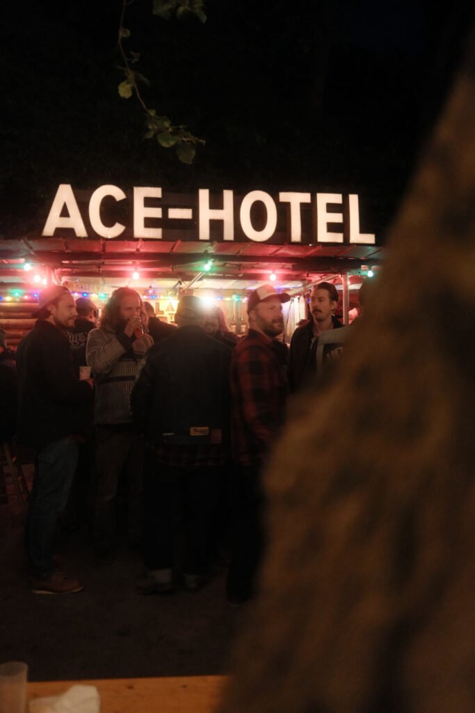 Eine Gruppe von Menschen steht und unterhält sich unter bunten Lichterketten vor einem Gebäude mit einem großen leuchtenden "ACE-HOTEL"-Schild bei Nacht. Ein unscharfes Objekt ist teilweise im Vordergrund rechts zu sehen.