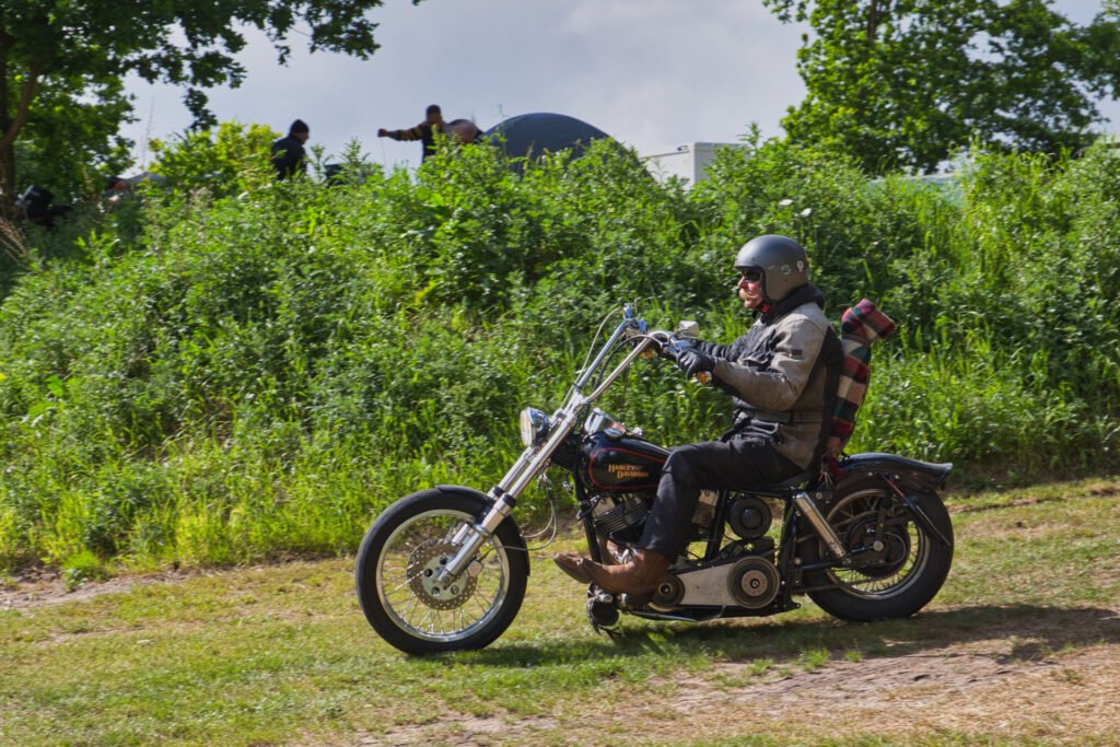 Eine Person, die einen Helm und eine Jacke trägt, fährt auf einem Motorrad im Vintage-Stil mit einer zusammengerollten Decke auf dem Rücken über einen grasbewachsenen Weg mit grünen Bäumen und Sträuchern im Hintergrund.