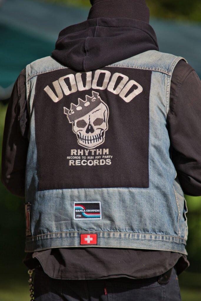 Eine Person trägt eine Jeansweste mit einem großen Aufnäher auf dem Rücken, der einen gekrönten Totenkopf und die Worte "VOODOO RHYTHM RECORDS-Records to ruin any party" zeigt. Auf der Weste befindet sich auch ein kleiner Aufnäher mit der Schweizer Flagge.
