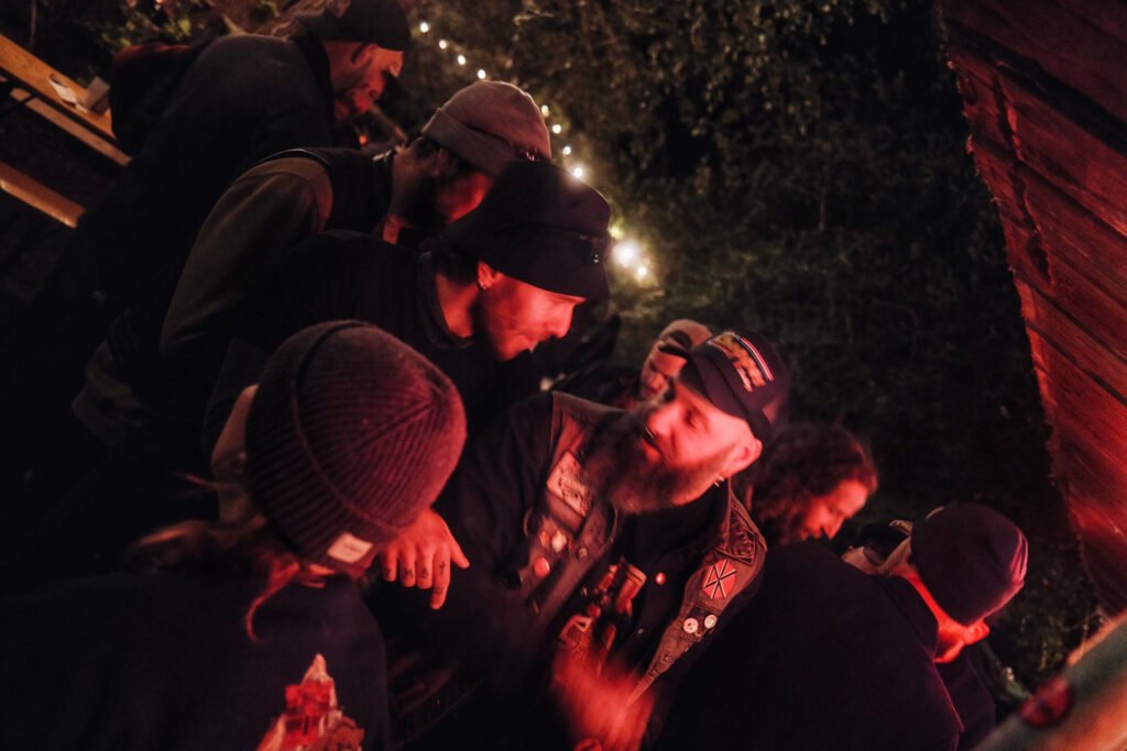 Eine Gruppe von Menschen mit Hüten und Jacken sitzt nachts unter Lichterketten eng beieinander und unterhält sich bei einer Veranstaltung im Freien. Die Szene ist warm beleuchtet und strahlt eine festliche Atmosphäre aus.