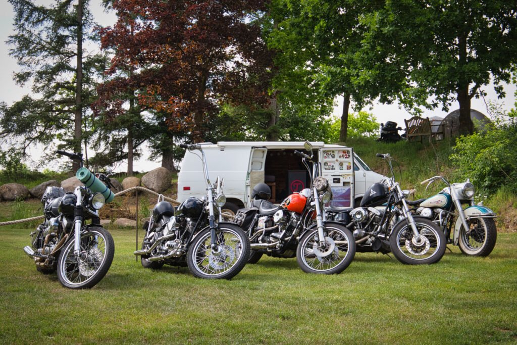 Fünf Oldtimer-Motorräder stehen auf einer Wiese vor einem weißen Lieferwagen mit offener Seitentür, umgeben von Bäumen und Felsen an einem sonnigen Tag. Auf einem der Motorräder ist eine Campingausrüstung zu sehen.