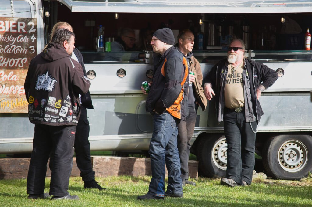 Eine Gruppe von Männern in Freizeit- und Bikerkleidung steht an einem sonnigen Tag vor einem Essens- oder Getränkewagen und unterhält sich. Ein Mann trägt eine Sonnenbrille und ein Harley-Davidson-Shirt. Die Szene findet im Freien auf einer Wiese statt.
