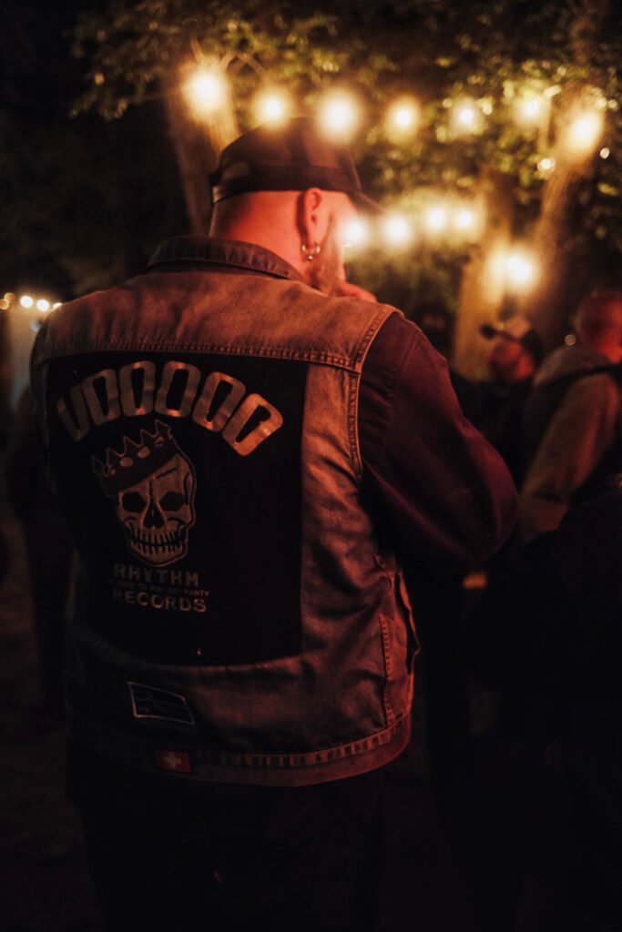 Eine Person, die eine Jeansweste mit einem Totenkopfmuster und "Voodoo Rhythm Records" auf dem Rücken trägt, steht nachts im Freien unter Lichterketten, umgeben von anderen Menschen.
