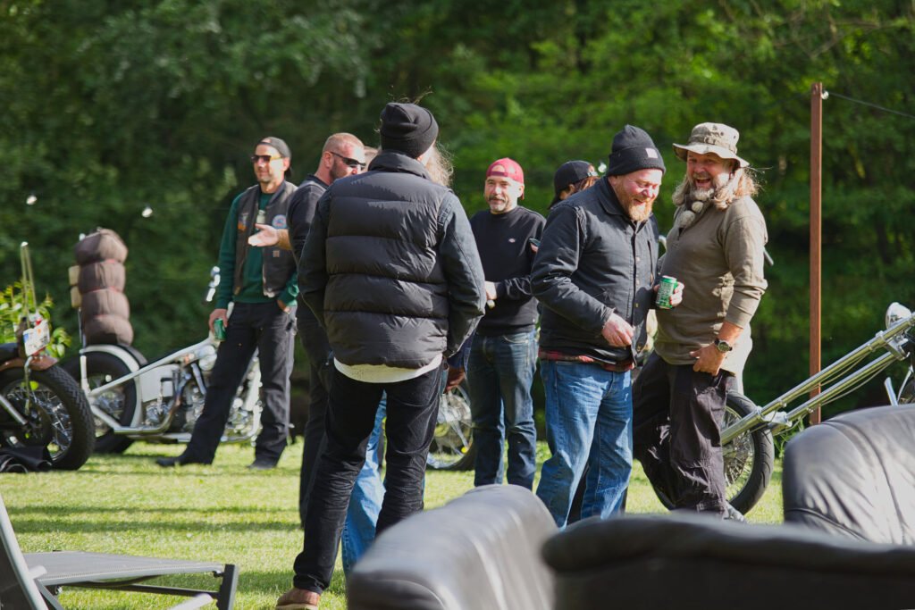 Eine Gruppe von Männern in legerer Outdoor-Kleidung und mit Mützen steht und unterhält sich in der Nähe von Motorrädern auf einer Wiese mit Bäumen im Hintergrund. Im Vordergrund sind ein paar Lederstühle zu sehen.