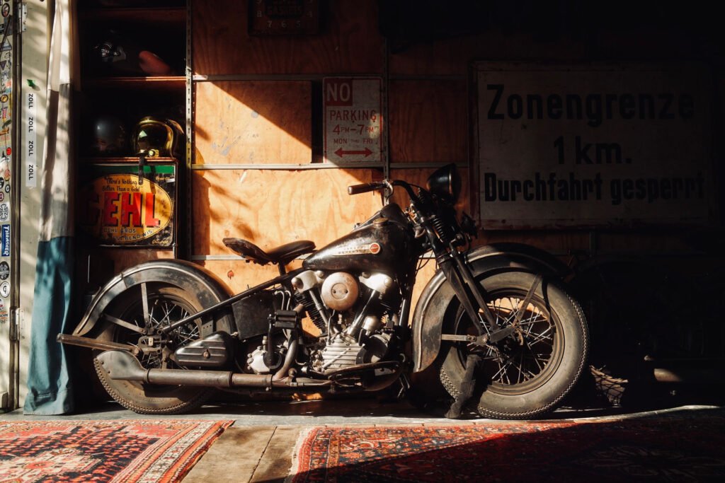 Ein schwarzes Oldtimer-Motorrad steht in einem Innenraum auf einem gemusterten Teppich, durch den warmes Sonnenlicht fällt. Dahinter stehen alte Holzregale, Helme und Retro-Schilder, darunter eines in deutscher Sprache und ein "Parkverbot"-Schild.