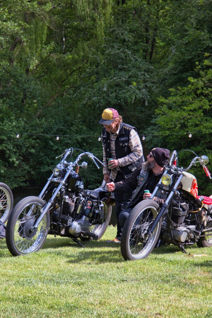 Zwei Personen mit Bärten und legerer Kleidung sitzen und stehen neben alten Motorrädern auf einer Wiese, unterhalten sich und halten Getränke in der Hand, mit grünen Bäumen im Hintergrund und Lichterketten darüber.