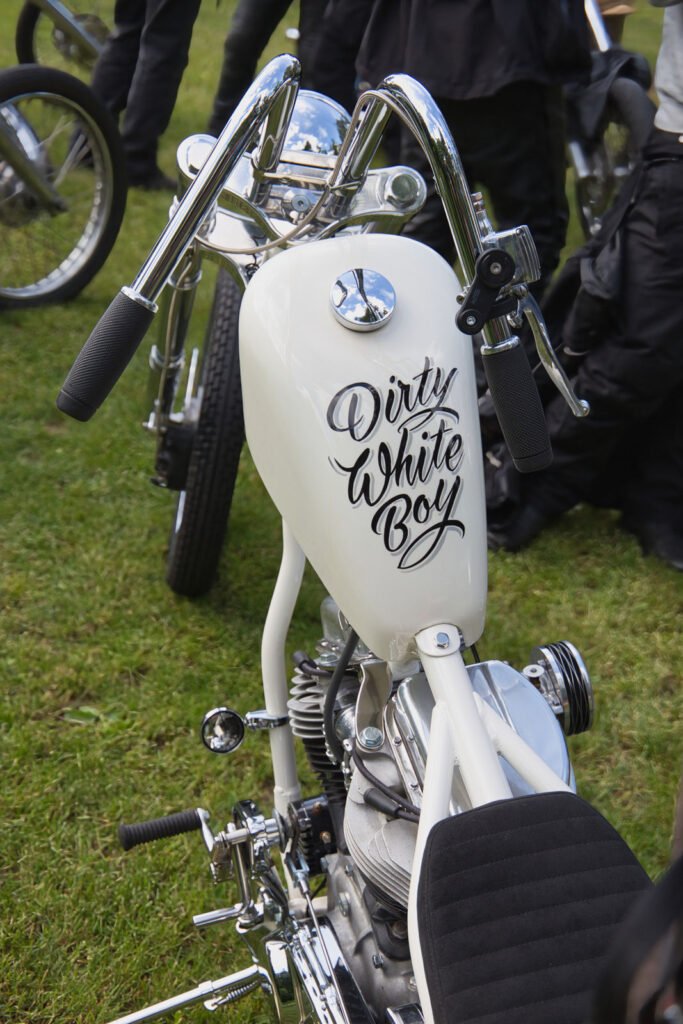 Nahaufnahme eines weißen Motorrads mit der Aufschrift "Dirty White Boy" in dekorativer Schrift auf dem Kraftstofftank, geparkt auf einer Wiese mit anderen Motorrädern und Menschen im Hintergrund.