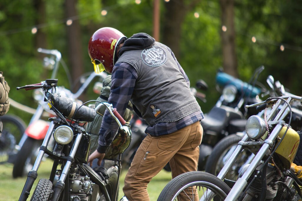 Eine Person, die einen roten Helm und eine karierte Jacke trägt, steht im Freien neben einem Oldtimer-Motorrad, mit mehreren anderen Motorrädern und Lichterketten im Hintergrund, umgeben von Grünpflanzen.
