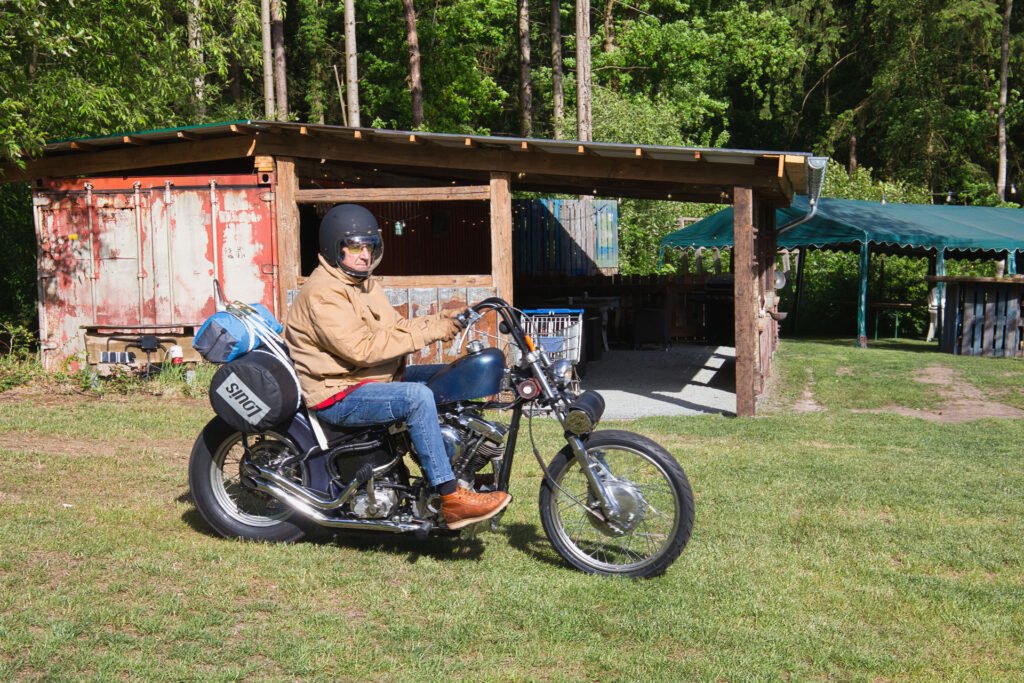 Eine Person mit Helm und Sonnenbrille sitzt auf einem schwarzen Motorrad mit einer "SINCE"-Tasche, das auf einer Wiese in der Nähe eines rustikalen Holzunterstands und eines verwitterten Schuppens geparkt ist, umgeben von Bäumen im Sonnenlicht.