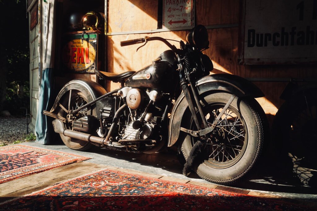 Ein schwarzes Oldtimer-Motorrad ist in einem Innenraum auf einem bunt gemusterten Teppich geparkt, das Sonnenlicht fällt ein und wirft Schatten; im Hintergrund sind alte Schilder und Dekoration zu sehen.