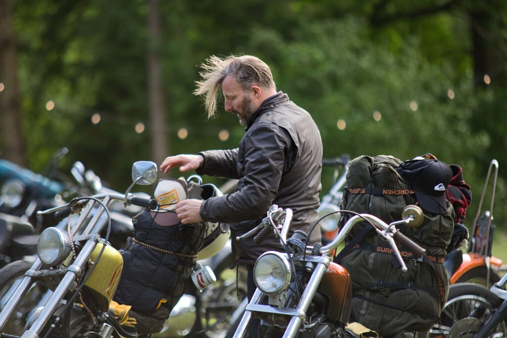 Eine Person mit vom Wind zerzaustem Haar steht im Freien zwischen geparkten Motorrädern und schnallt einen großen Rucksack auf eines der Räder. Im unscharfen Hintergrund sind Bäume und Lichterketten zu sehen.