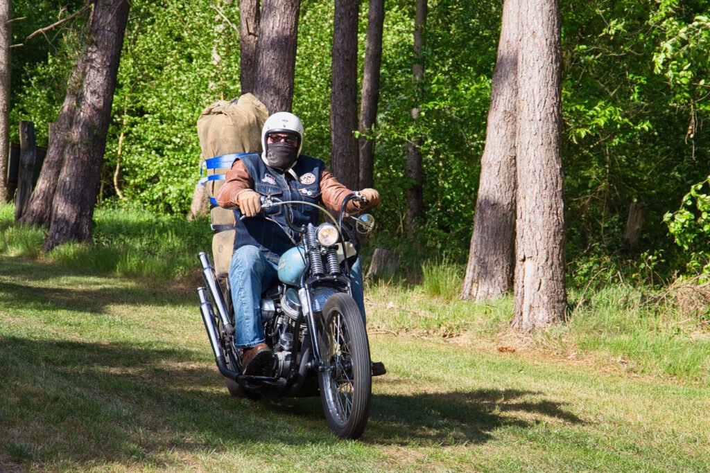 Eine Person, die einen Helm und eine Sonnenbrille trägt, fährt auf einem Motorrad durch einen sonnenbeschienenen Wald. Hinten auf dem Motorrad ist ein großer Rucksack angeschnallt. Der Boden ist grasbewachsen, und hohe Bäume umgeben die Szene.