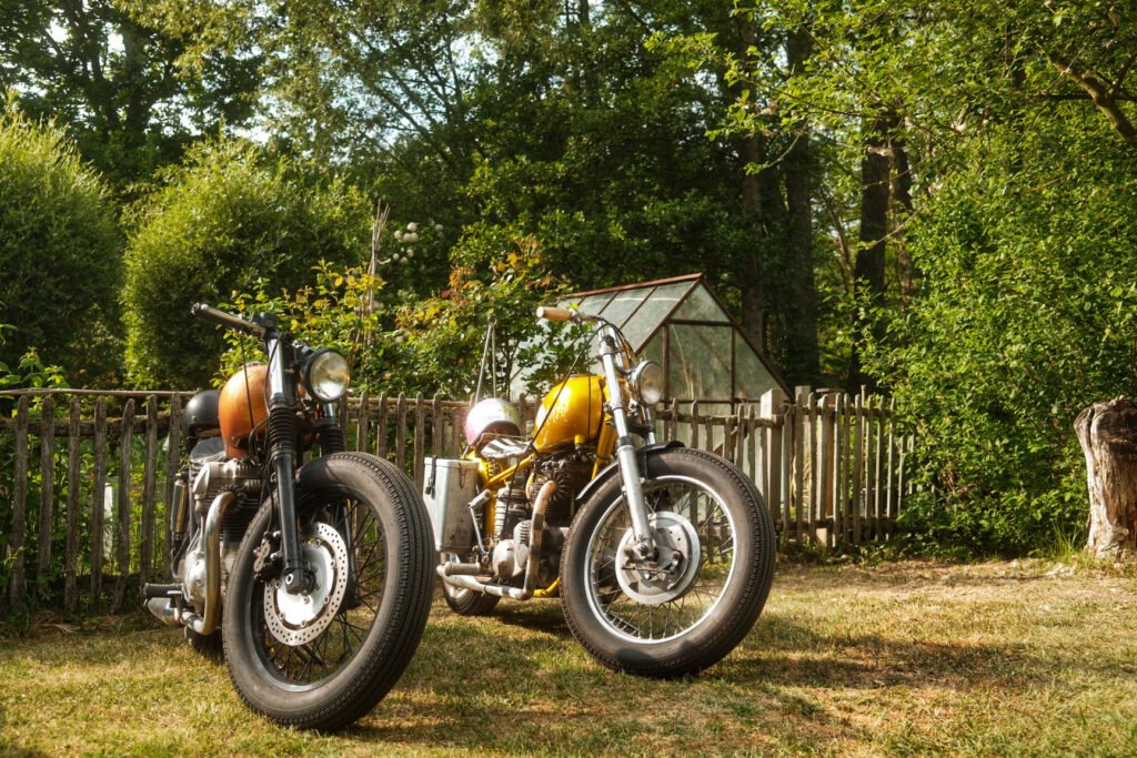 Zwei Oldtimer-Motorräder mit orangefarbenen und gelben Tanks sind auf trockenem Gras vor einem Holzzaun geparkt, mit einem kleinen Glasgewächshaus und üppigen grünen Bäumen im Hintergrund.