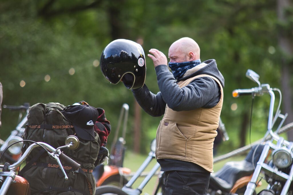 Ein Mann mit hellbrauner Weste und blauer Bandana-Maske steht im Freien und hält einen schwarzen Motorradhelm an seinen Kopf. In der Nähe stehen mehrere Motorräder und ein Rucksack, im Hintergrund sind grüne Bäume zu sehen.