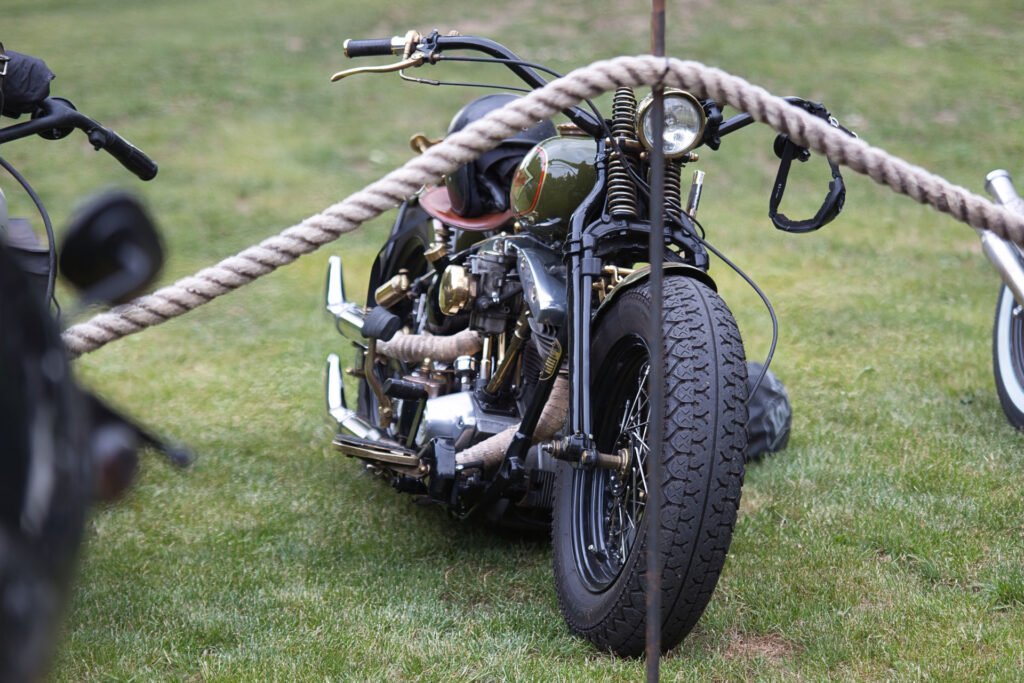 Ein speziell angefertigtes Oldtimer-Motorrad mit schwarzen Reifen, verchromten Teilen und Ledersitz ist hinter einer dicken Seilabsperrung im Gras geparkt. Der Schauplatz scheint eine Veranstaltung im Freien zu sein.