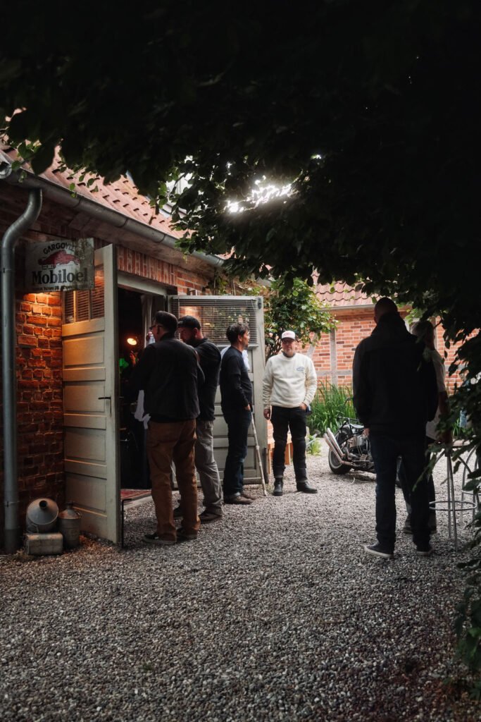 Mehrere Personen stehen und unterhalten sich vor einem Backsteingebäude mit offener Tür, umgeben von Grün und Kies. Über dem Eingang hängt ein Schild mit der Aufschrift "Mobile", und im Hintergrund ist ein Fahrrad geparkt.