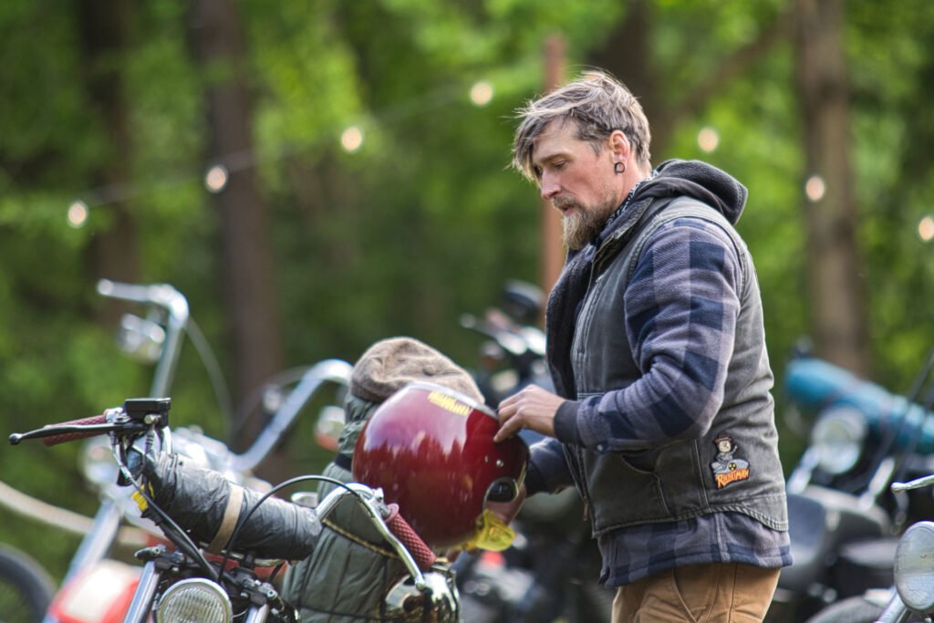 Ein Mann mit Bart und karierter Jacke steht im Freien neben einem Motorrad und trägt einen roten Helm. Im Hintergrund sind mehrere Motorräder geparkt, und es sind grüne Bäume und Lichterketten zu sehen.